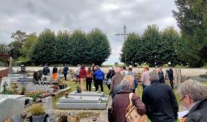 Cimetières, chaque tombe est entourée de pelouse, un groupe d'une 30taine de personnes circulent entre les tombes gris clair. Des arbre derrière le muret du cimetière au fond. et une croix domine le groupe et les tombes juste devant ce mur.