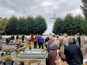 Cimetières, chaque tombe est entourée de pelouse, un groupe d'une 30taine de personnes circulent entre les tombes gris clair. Des arbre derrière le muret du cimetière au fond. et une croix domine le groupe et les tombes juste devant ce mur.