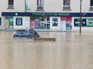 Une place inondée, dans le fond une pharmacie, une voiture au milieu dans l'eau.