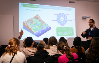Un groupe d'élèves écoute l'intervention d'un agent du Département. Un diaporama présente le grand cycle de l'eau. L'animateur est en train de donner la parole à un élève qui lève le doigt.