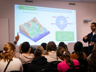 Un groupe d'élèves écoute l'intervention d'un agent du Département. Un diaporama présente le grand cycle de l'eau. L'animateur est en train de donner la parole à un élève qui lève le doigt.
