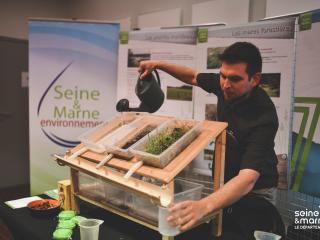 Présentation d'une maquette expliquant le ruissellement des eaux pluviales sur un stand de Seine-et-Marne Environnement. La pluie est simulée par un arrosoir sur différents supports : herbe, terre, sol imperméable