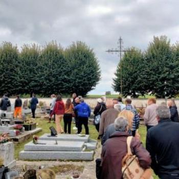 Cimetières, chaque tombe est entourée de pelouse, un groupe d'une 30taine de personnes circulent entre les tombes gris clair. Des arbre derrière le muret du cimetière au fond. et une croix domine le groupe et les tombes juste devant ce mur.