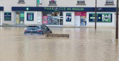 Une place inondée, dans le fond une pharmacie, une voiture au milieu dans l'eau.