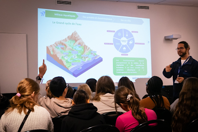 Un groupe d'élèves écoute l'intervention d'un agent du Département. Un diaporama présente le grand cycle de l'eau. L'animateur est en train de donner la parole à un élève qui lève le doigt.
