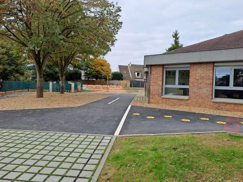 Vue de la cour d'école désimperméabilisée avec au premier plan un espace enherbé, un espace en pavés à joints enherbés, en second plan des cheminements en bitume ornés d'empruntes animales peintes, et en arrière plan des arbres au sein d'un espace perméable en copeaux de bois
