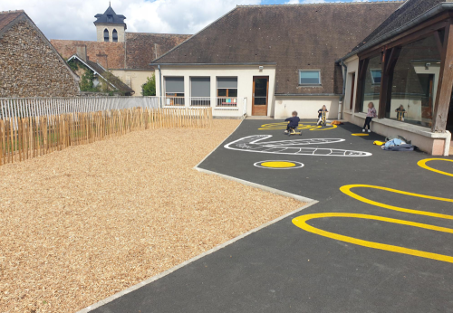 Cour désimperméabilisée avec à gauche un ilot d'infiltration paillé qui sera végétalisé. A droite, des enfants roulent avec des tricycles sur des parcours peints sur une partie en bitume/