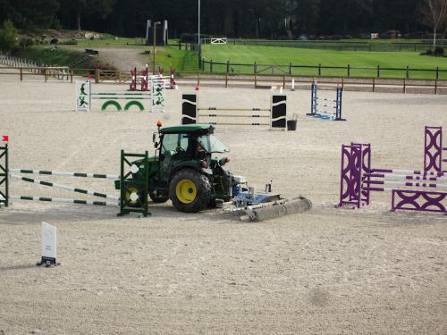Dans un carrière équestre en sable, un tracteur tire un module de désherbage mécanique, entre les haies de saut d'obstacle