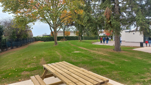 Cour d'école avec au premier plan une table type "pique-nique" en bois au sein d'un espace vert enherbé avec de grands arbres