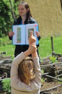 Un enfant lève le doigt face à une animatrice montre un document. La scène se passe en extérieur près d'un potager.